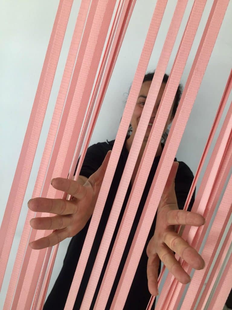 une femme cachée derrière des élastique tendu de couleur rose sort ses mains à travers